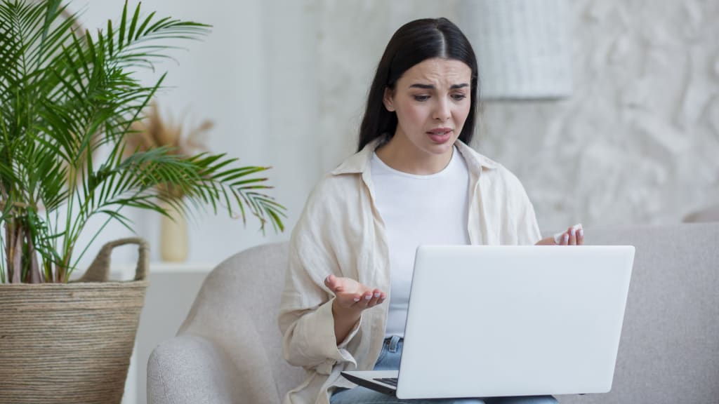 A woman sitting on a couch looks frustrated while using a laptop, possibly searching for answers to questions like "How Much Does BetterHelp Pay Therapists?